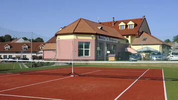 A tennis court with a net, surrounded by buildings and parked cars.