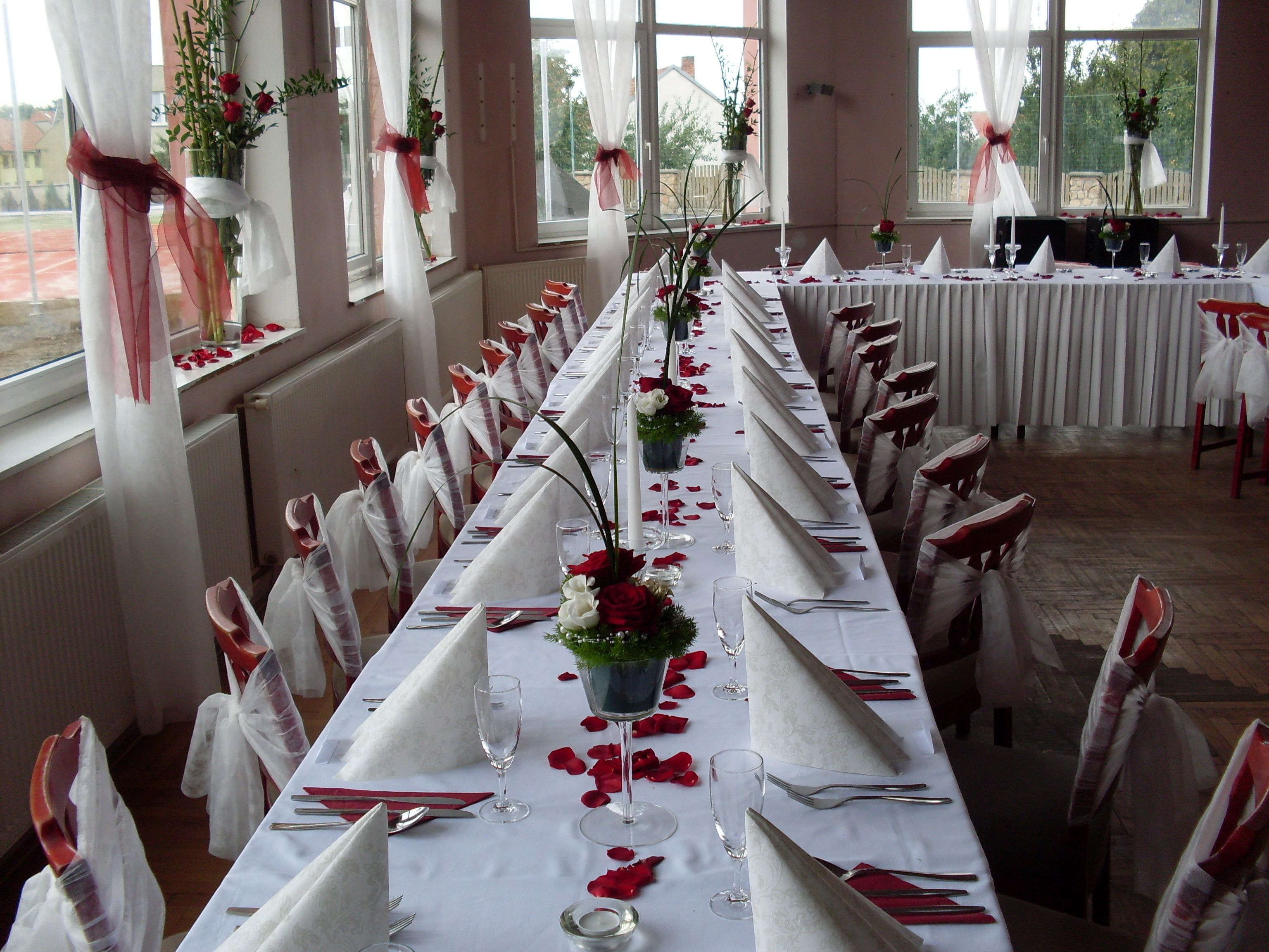 Une longue table de banquet, dressée pour un événement formel, avec des nappes blanches, des pétales de roses rouges et des chaises blanches et rouges.