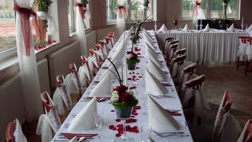 Une longue table de banquet, dressée pour un événement formel, avec des nappes blanches, des pétales de roses rouges et des chaises blanches et rouges.