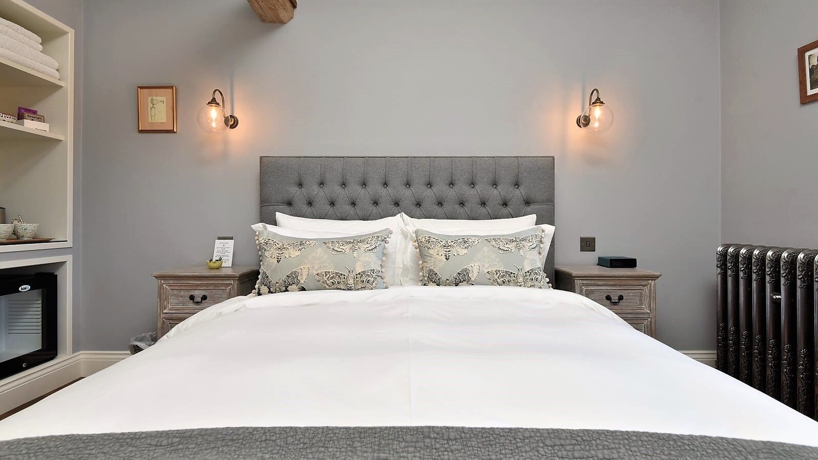 Una cama bien hecha con cabecera gris, dos mesitas de noche con lámparas, un radiador y un estante con toallas y un cuenco.