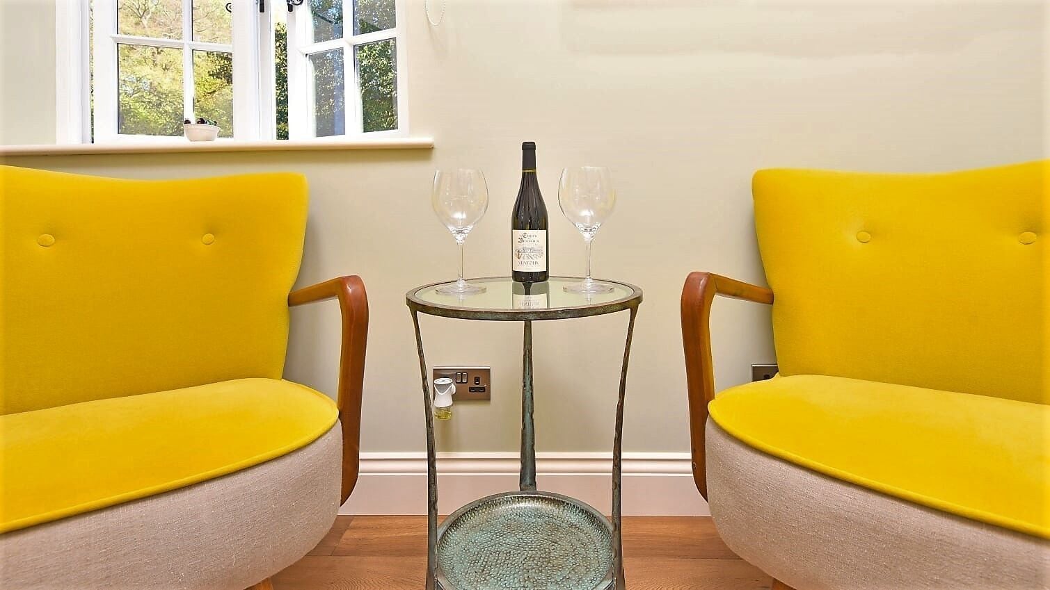 A room with two yellow chairs, a small round table with a bottle of wine and two glasses, and a window with a view of greenery.