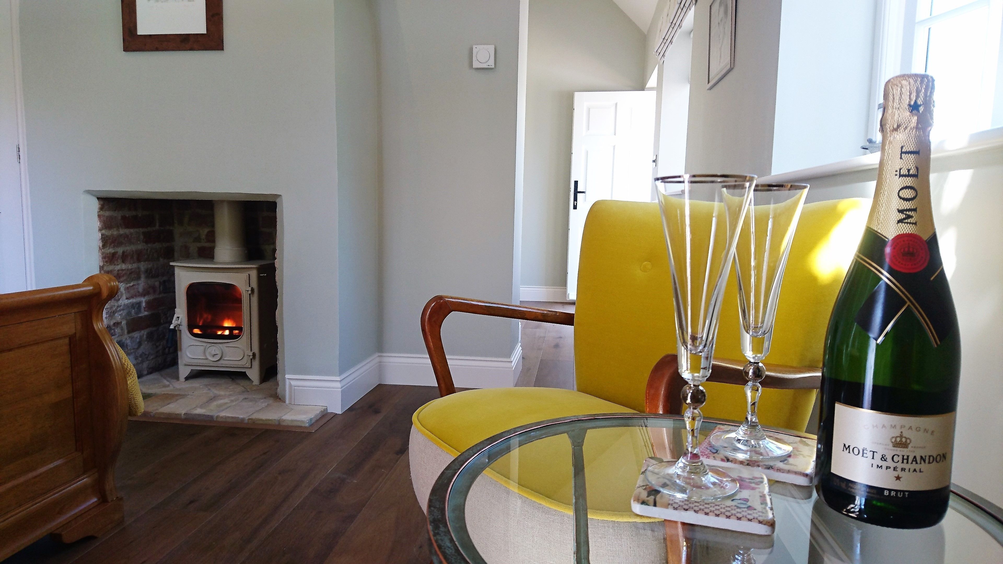 A room with a fireplace, a yellow armchair, a glass table with a bottle of Moët & Chandon champagne, and a wooden chair.