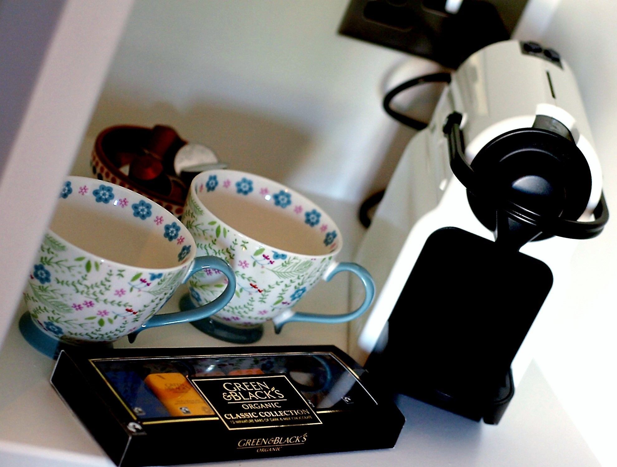 A coffee maker, two floral-patterned cups, and a box of Green & Black's organic coffee.