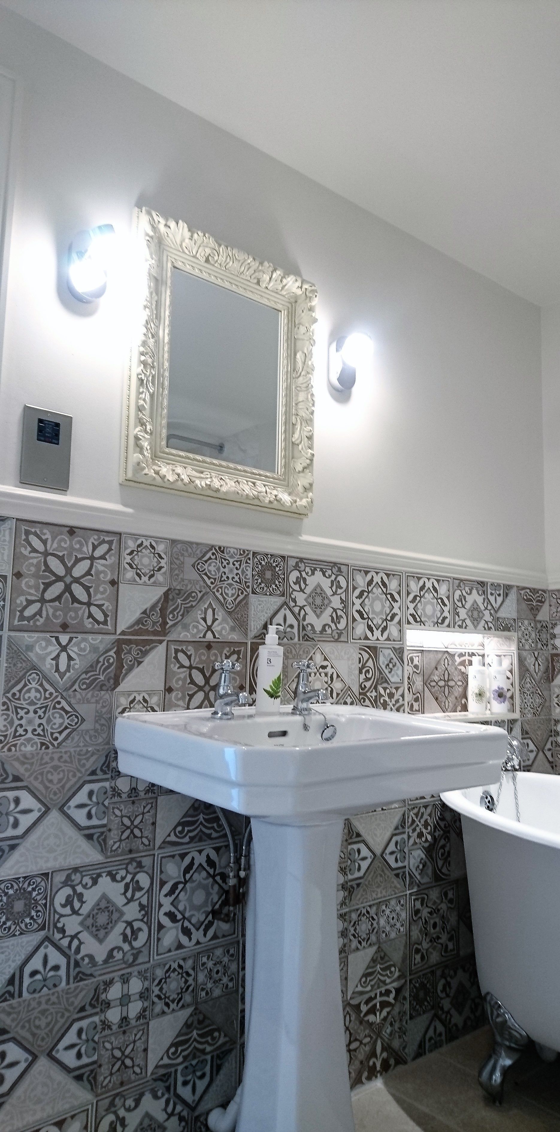 A bathroom with a white pedestal sink, a large ornate mirror, and patterned tiles on the walls.
