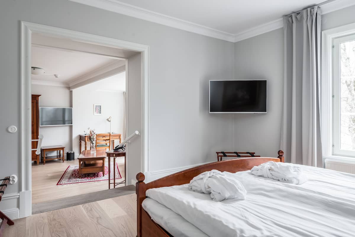 A hotel room with a bed, a TV mounted on the wall, a wooden chair, a small table, and a window with curtains.