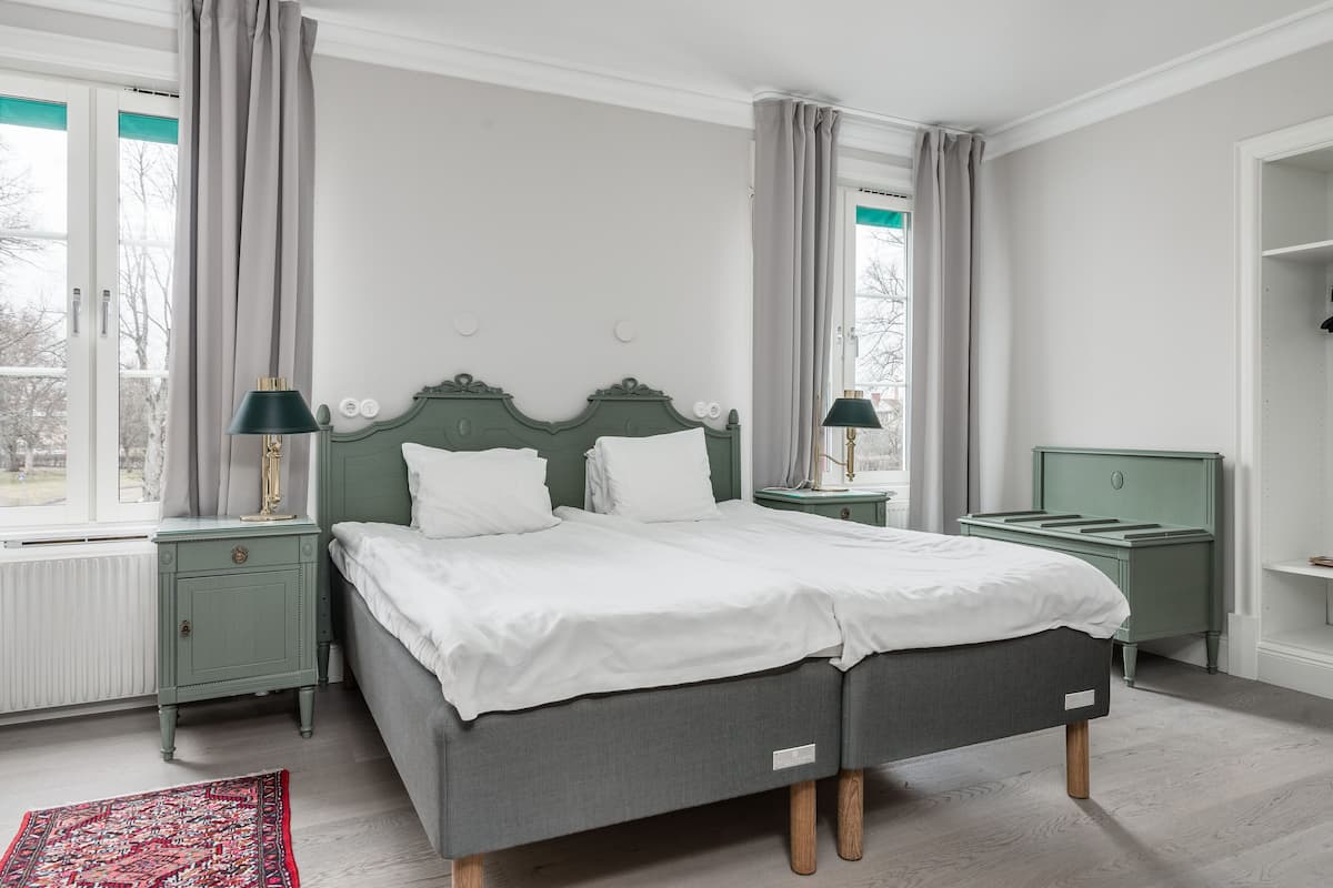 A double bed with a green headboard, two bedside tables with lamps, a red patterned rug, and a view of trees through a window.