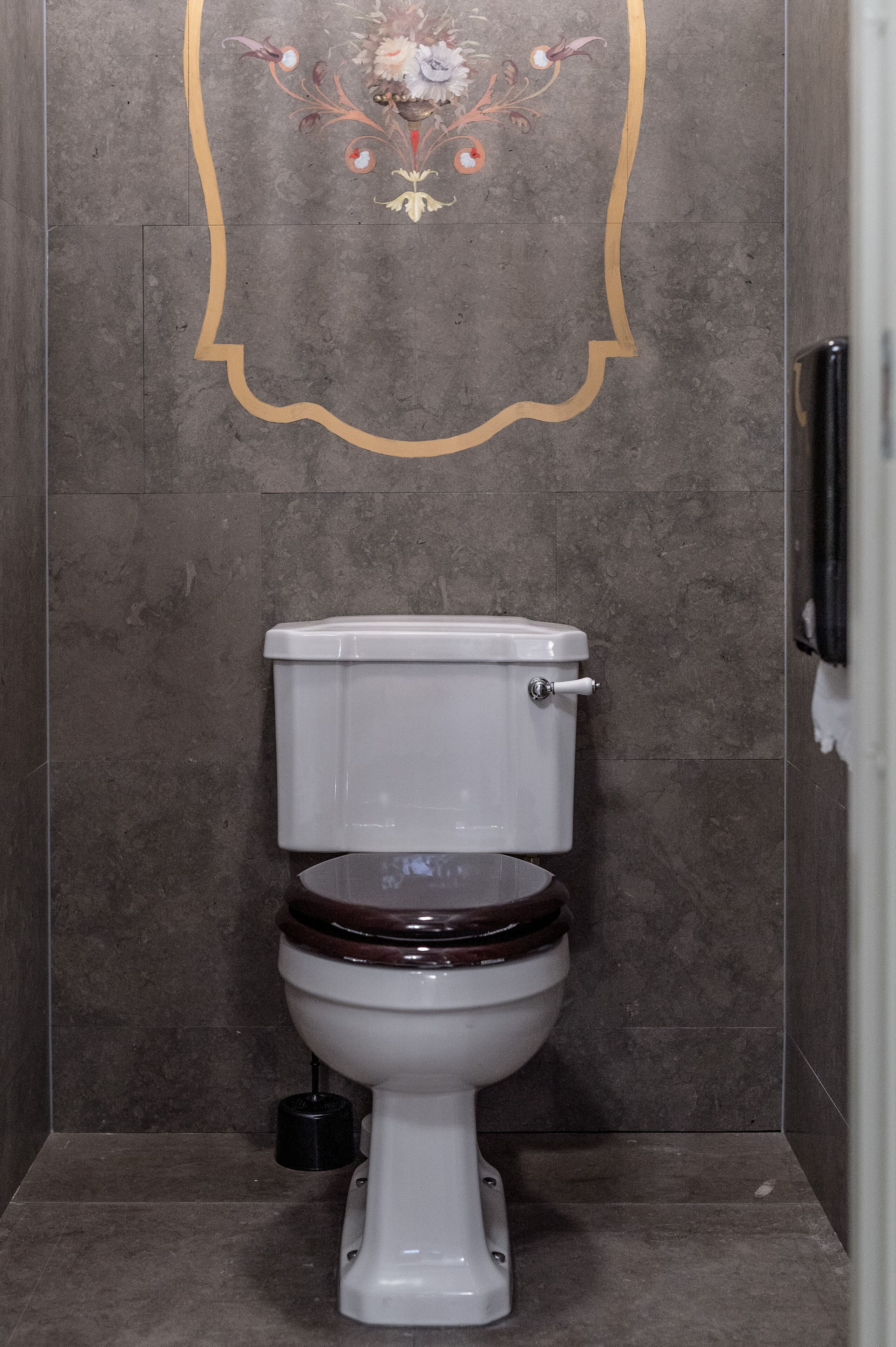 Un inodoro con asiento y tapa de madera, un escobilla negra y un diseño decorativo en la pared.