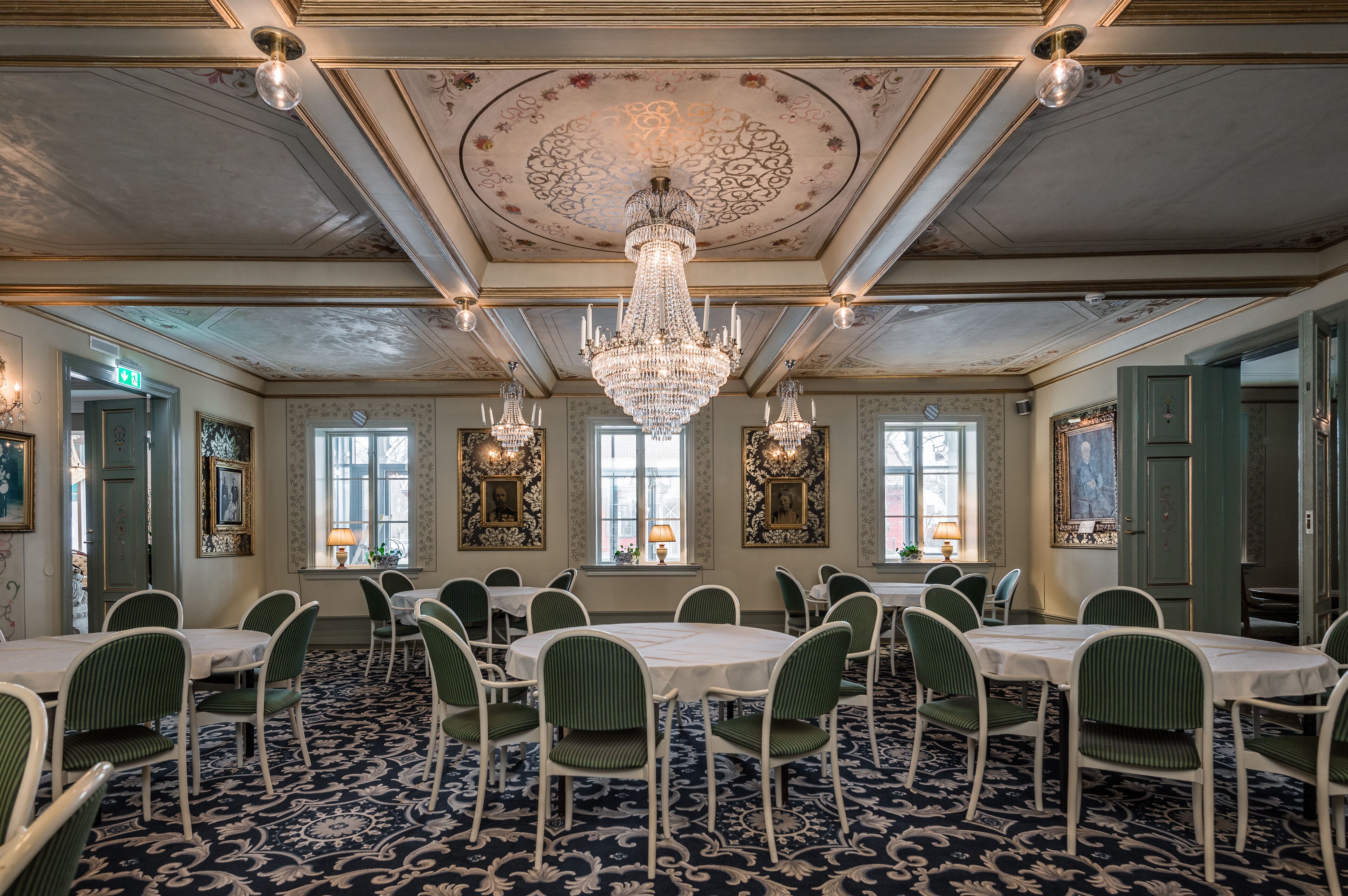 Une salle à manger élégante, dotée de lustres, de fauteuils recouverts d’un tissu vert et d’un tapis à motifs.