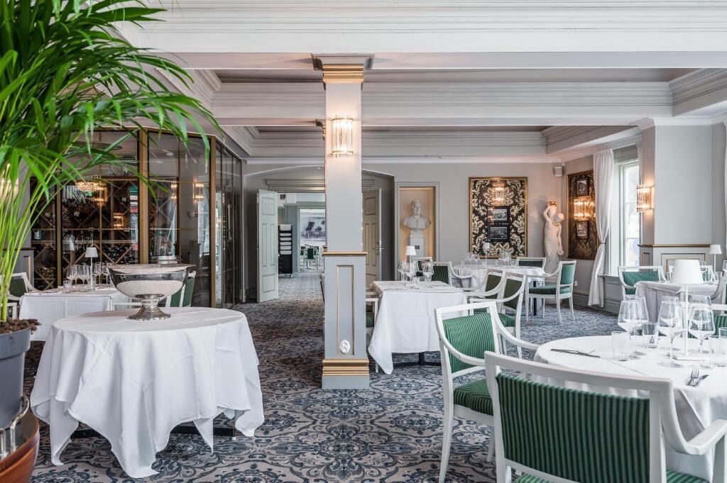 A restaurant with round tables, white tablecloths, and green chairs. There are potted plants and a patterned carpet.