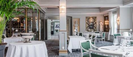 A restaurant with round tables, white tablecloths, and green chairs. There are potted plants and a patterned carpet.