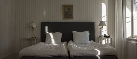 A double bed with white linens, two bedside tables with lamps, a framed picture on the wall, and a window with curtains.