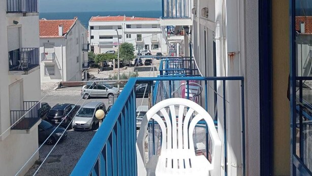 Un balcón con una silla de plástico blanco, barandilla azul y vista al mar y edificios.