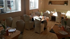 A dining area with multiple tables and chairs, large windows with curtains, and a brick wall with a shelf. - Hotel Garni Haus Sonneneck (Thale)