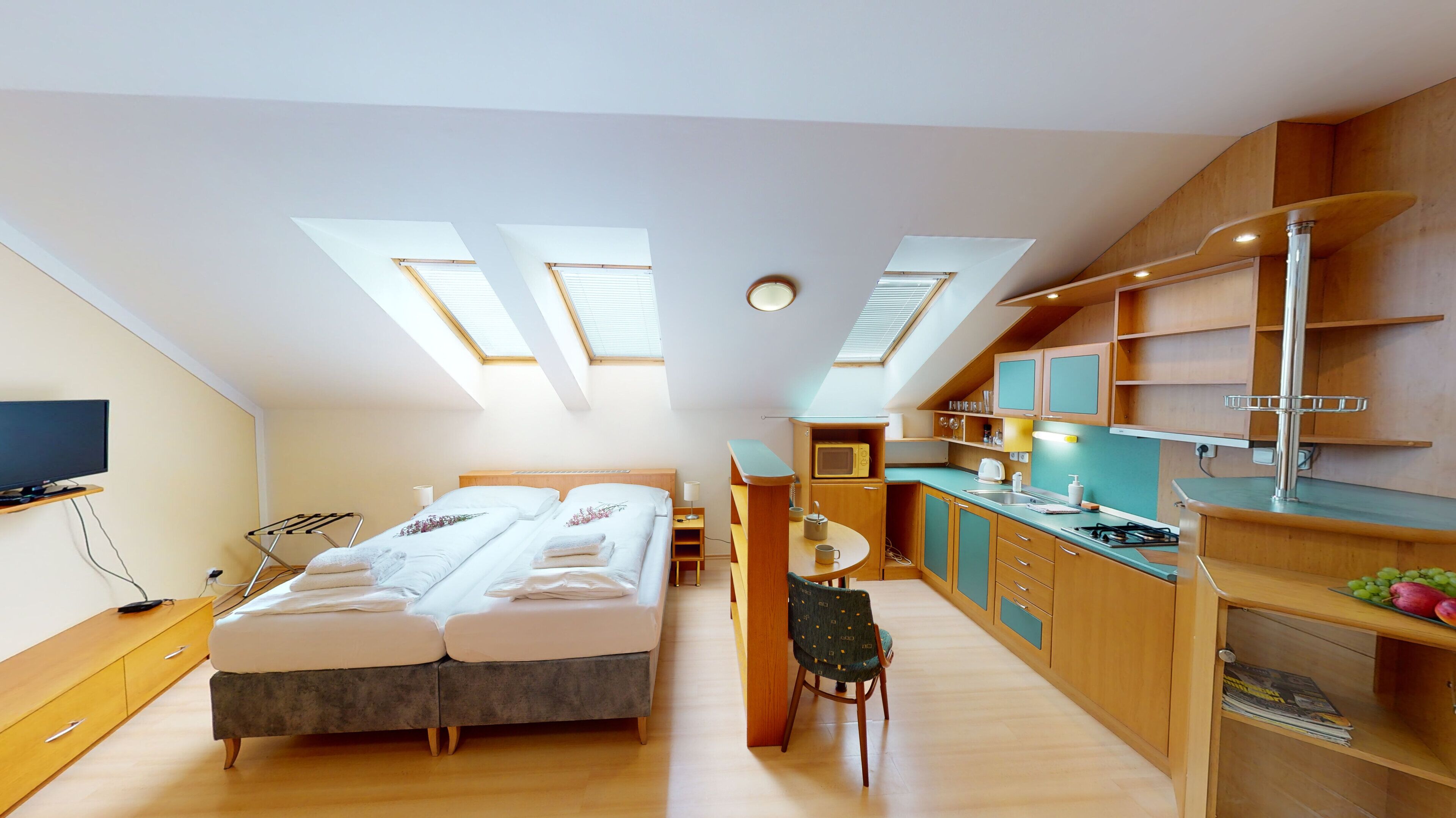 A compact hotel room with a bed, a small table, a chair, a TV, and a kitchenette.
