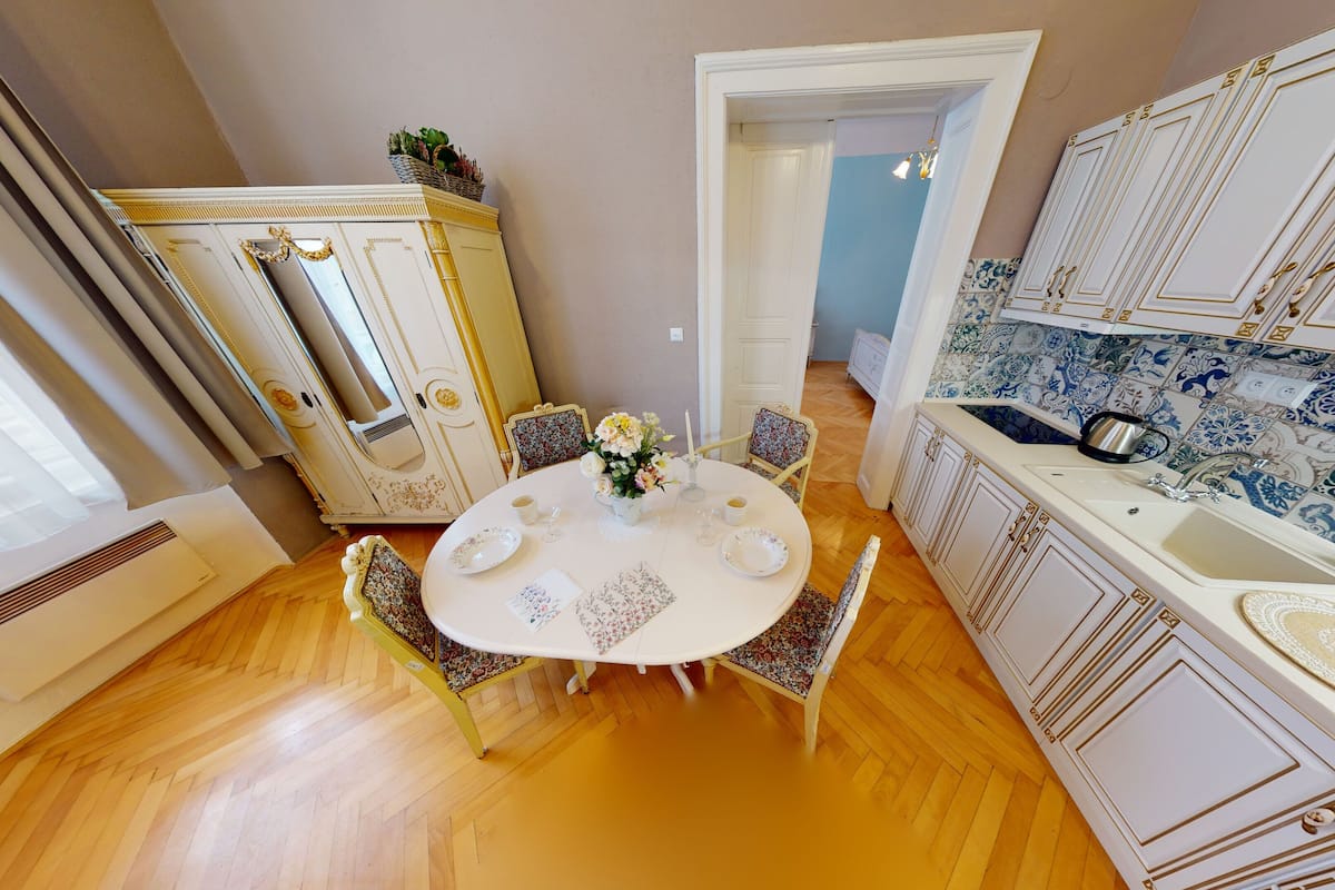 A dining area with a round table set for four, surrounded by ornate chairs. A white cabinet with gold detailing is visible in the background, and a kitchen area with blue and white patterned tiles is to the right.