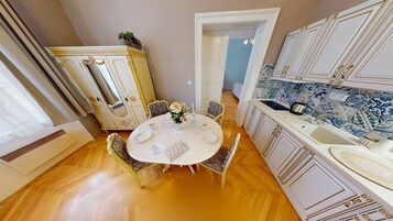 Un comedor con una mesa redonda dispuesta para cuatro, rodeada de sillas ornamentadas. Se observa un aparador blanco con detalles dorados al fondo, y un área de cocina con baldosas azules y blancas a la derecha.
