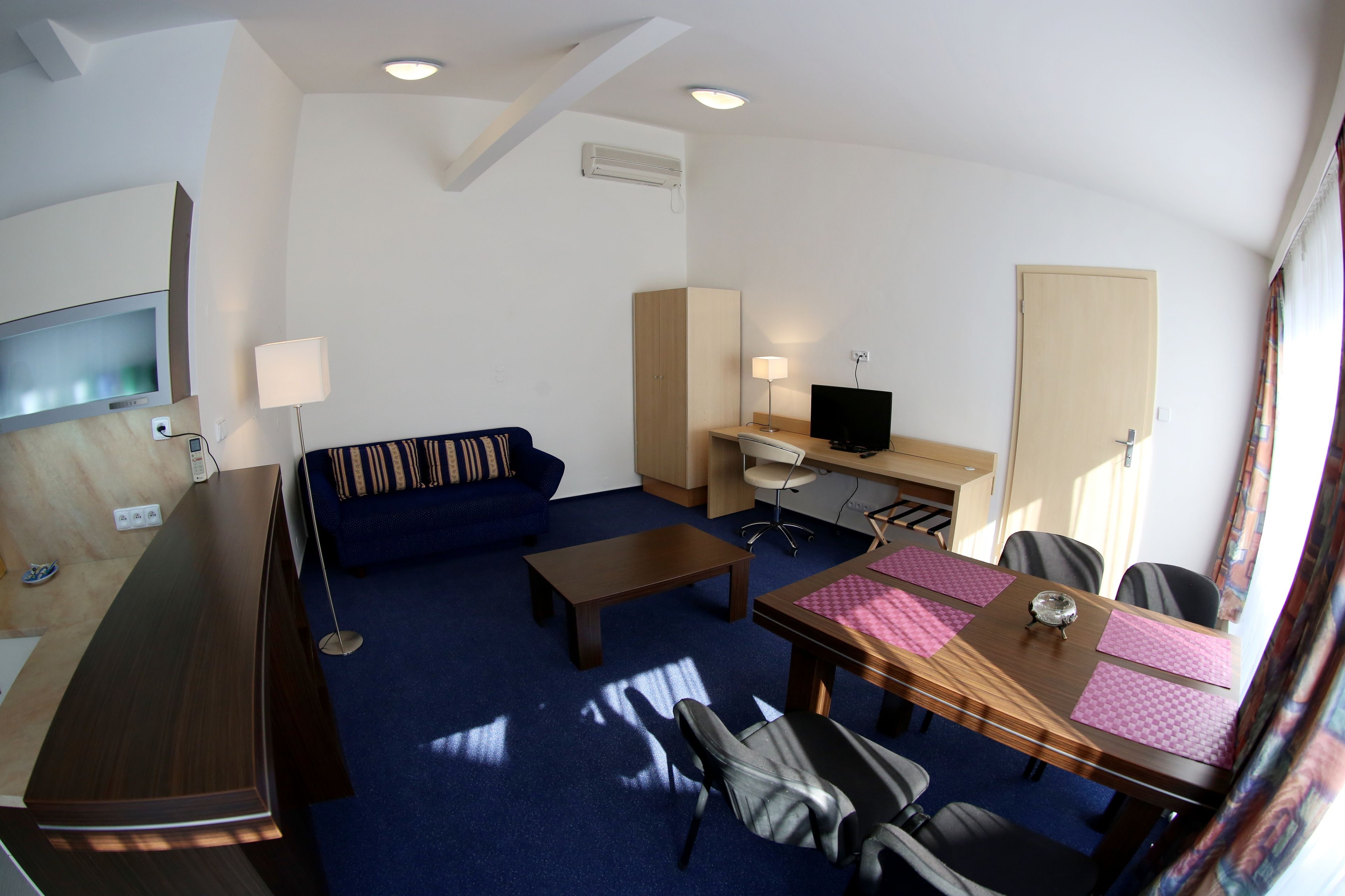 A hotel room with a long wooden desk, a blue sofa, a small table, a TV, and a desk lamp.