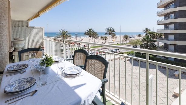 A balcony with a table set for dining, overlooking a beach with palm trees and a clear sky. - Ocean view condo located in front of the beach with sunny terrace - only families (Playa de Gandía)