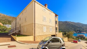 A small, two-story stone building with a balcony, a small car parked in front, and a scenic view of a lake and mountains in the background. - New brand apartment with sea view  (Dubrovnik)
