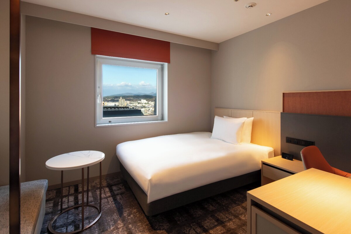 A hotel room with a bed, a small round table, a desk, and a view of a cityscape through a window.