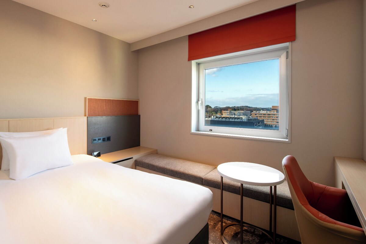 A hotel room with a large bed, a small table, a chair, and a window with a city view.