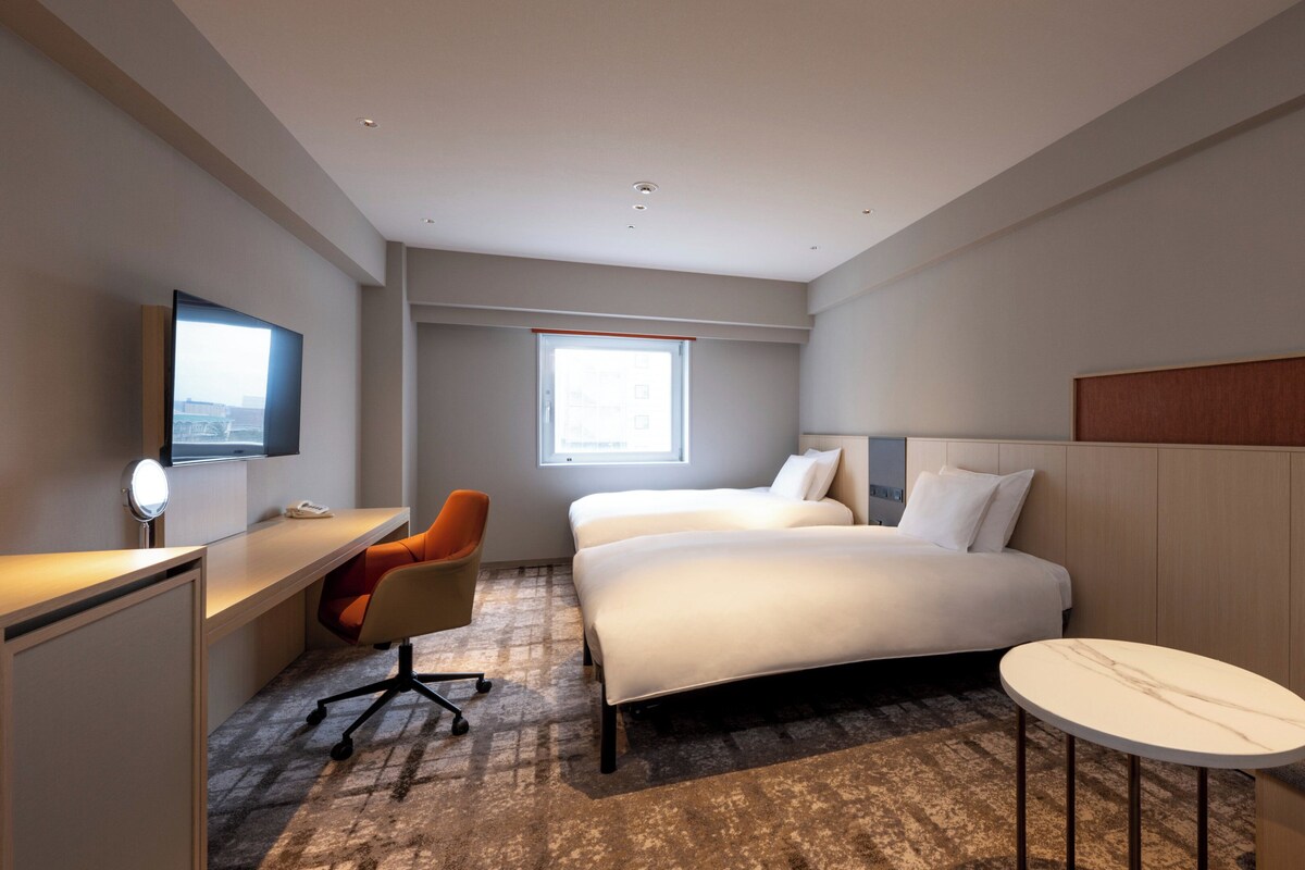 A hotel room with a bed, a desk, an orange chair, a small round table, and a TV mounted on the wall.