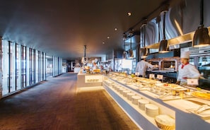 Una cocina moderna con chefs preparando comida, una barra de buffet con platos y cubiertos, y amplios ventanales a lo largo del pasillo.