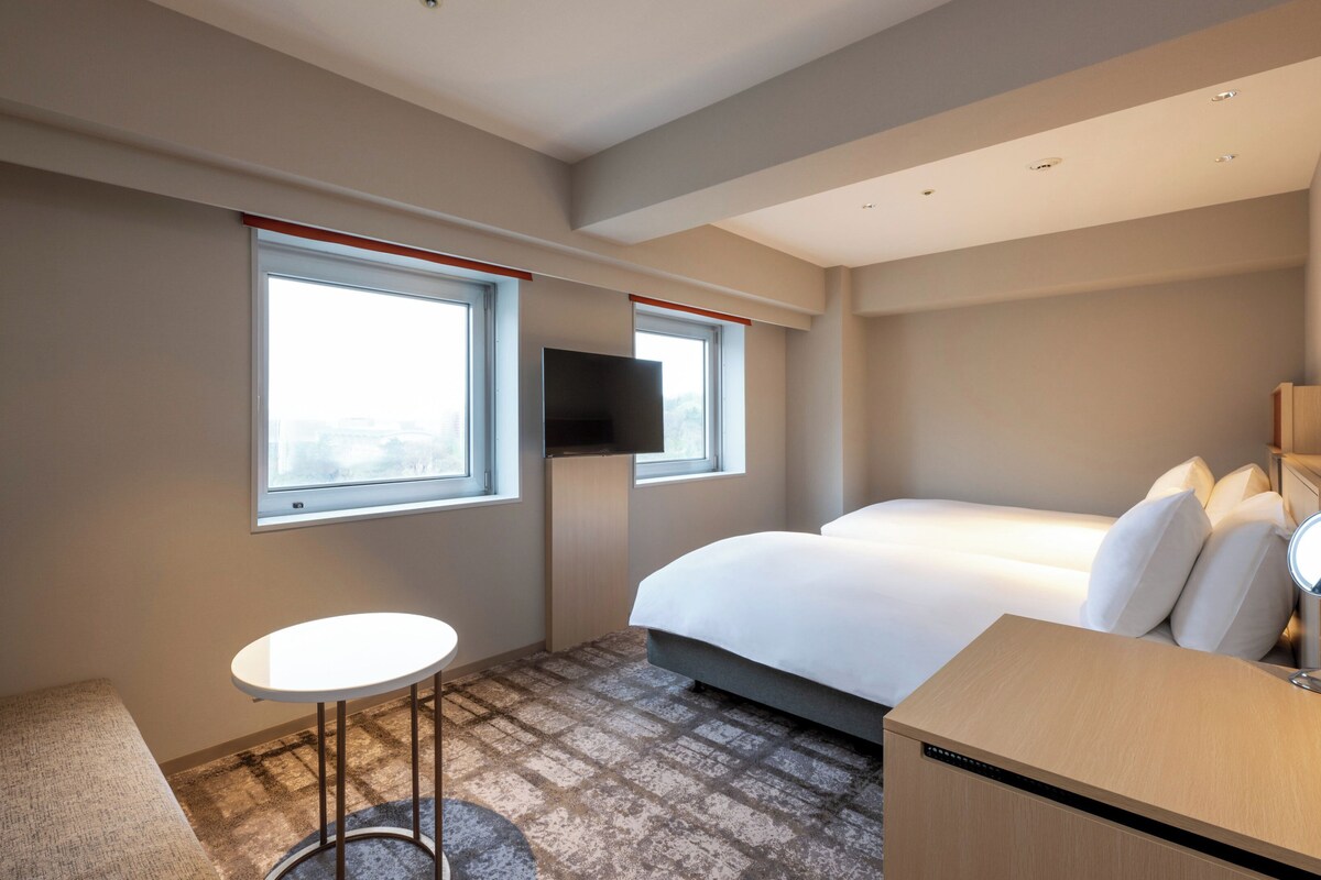 A hotel room with a bed, a small table, a TV mounted on the wall, and a window with a view.