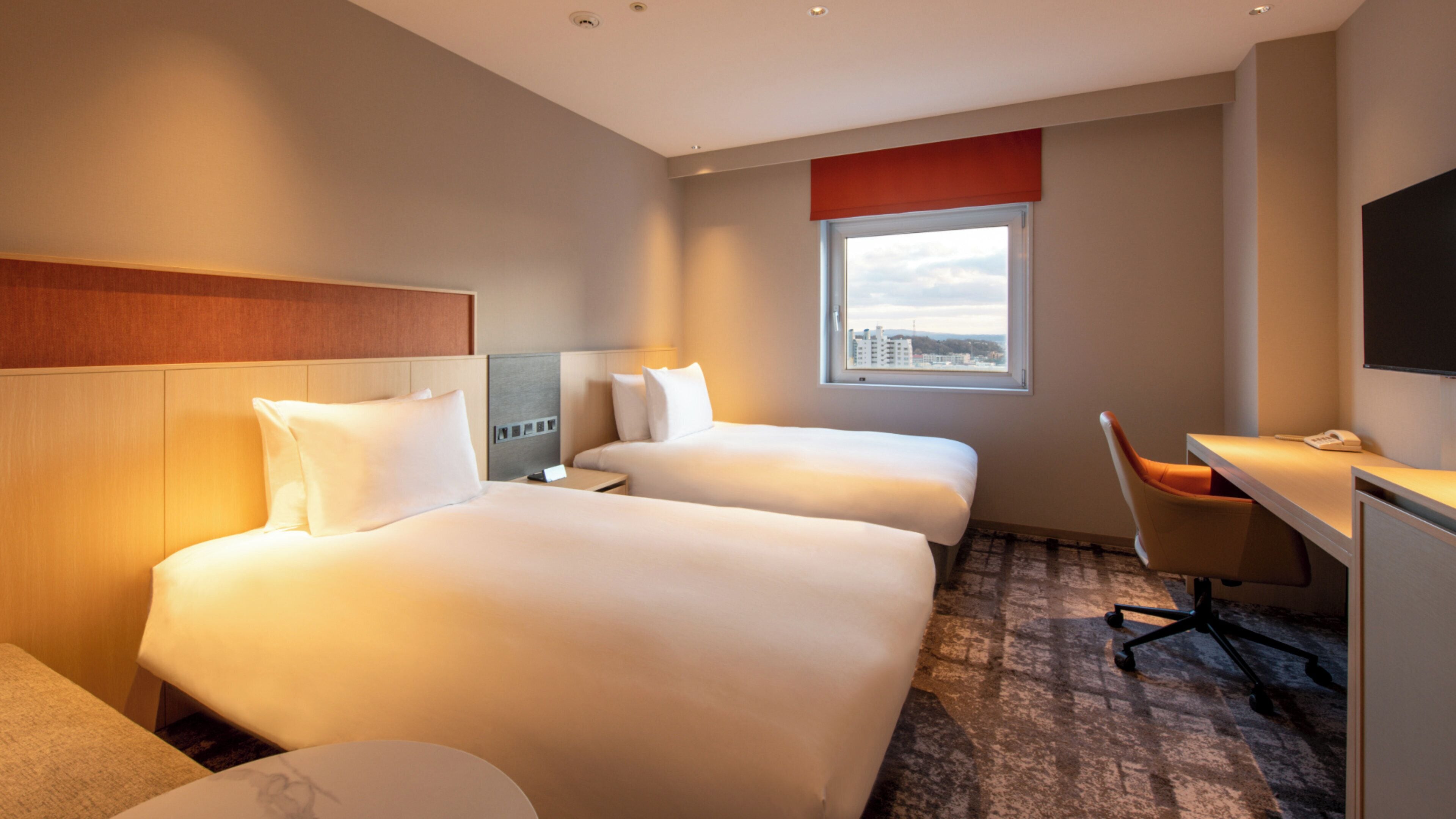 A hotel room with two beds, a desk, a chair, and a window with a city view.