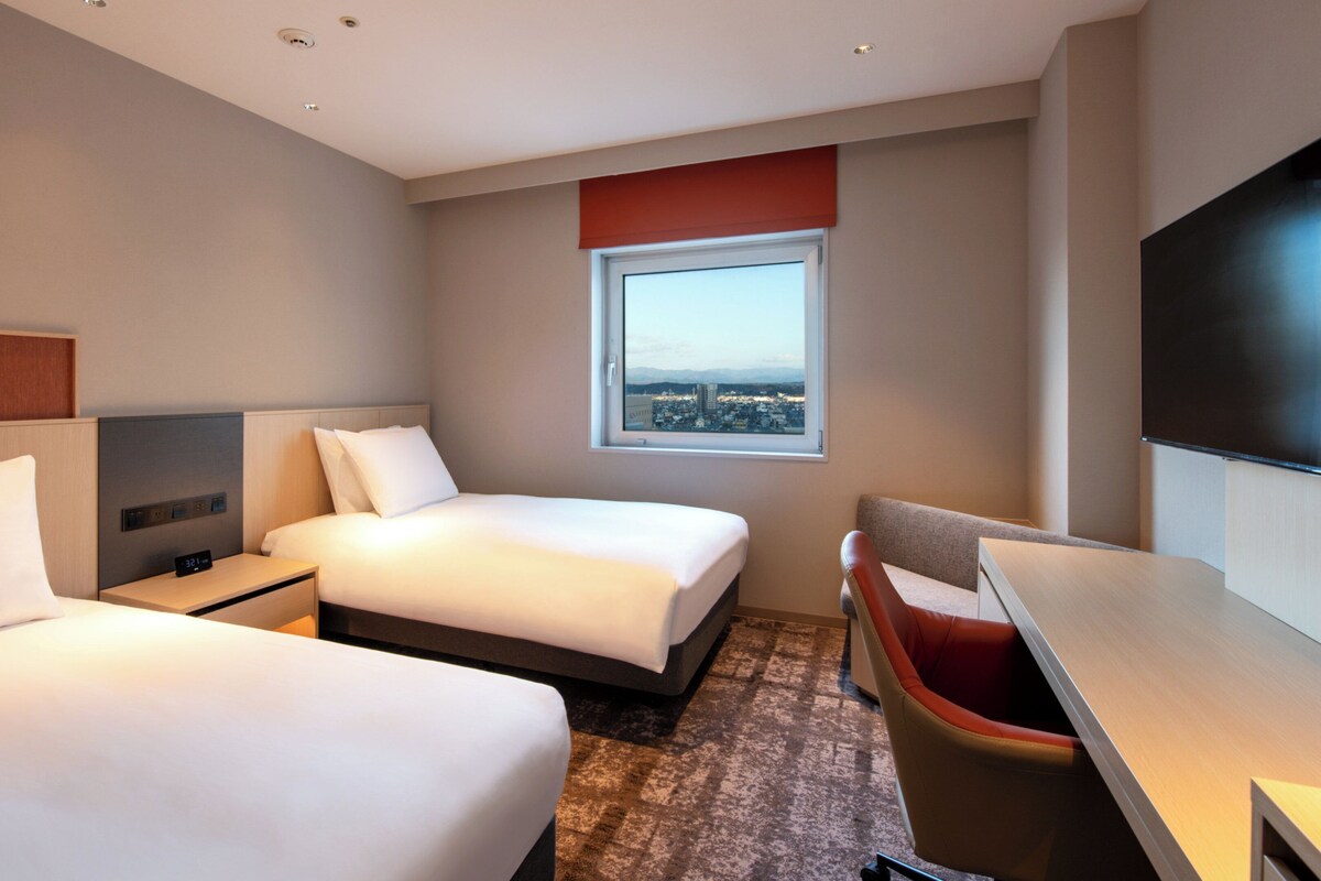 A hotel room with two beds, a desk, a chair, a TV, and a window with a city view.