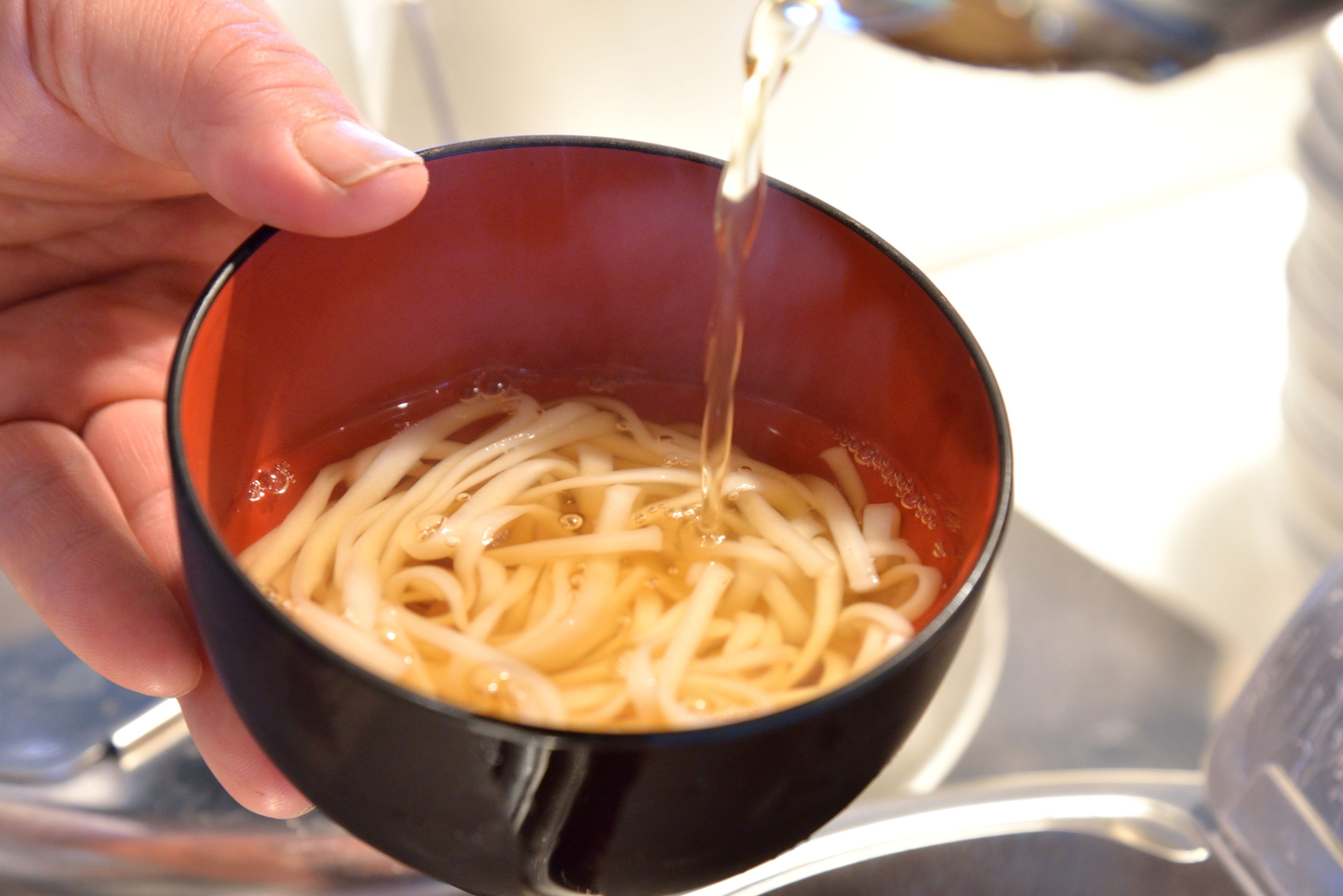 Una mano sosteniendo un tazón de fideos, con un líquido transparente que se vierte en su interior.