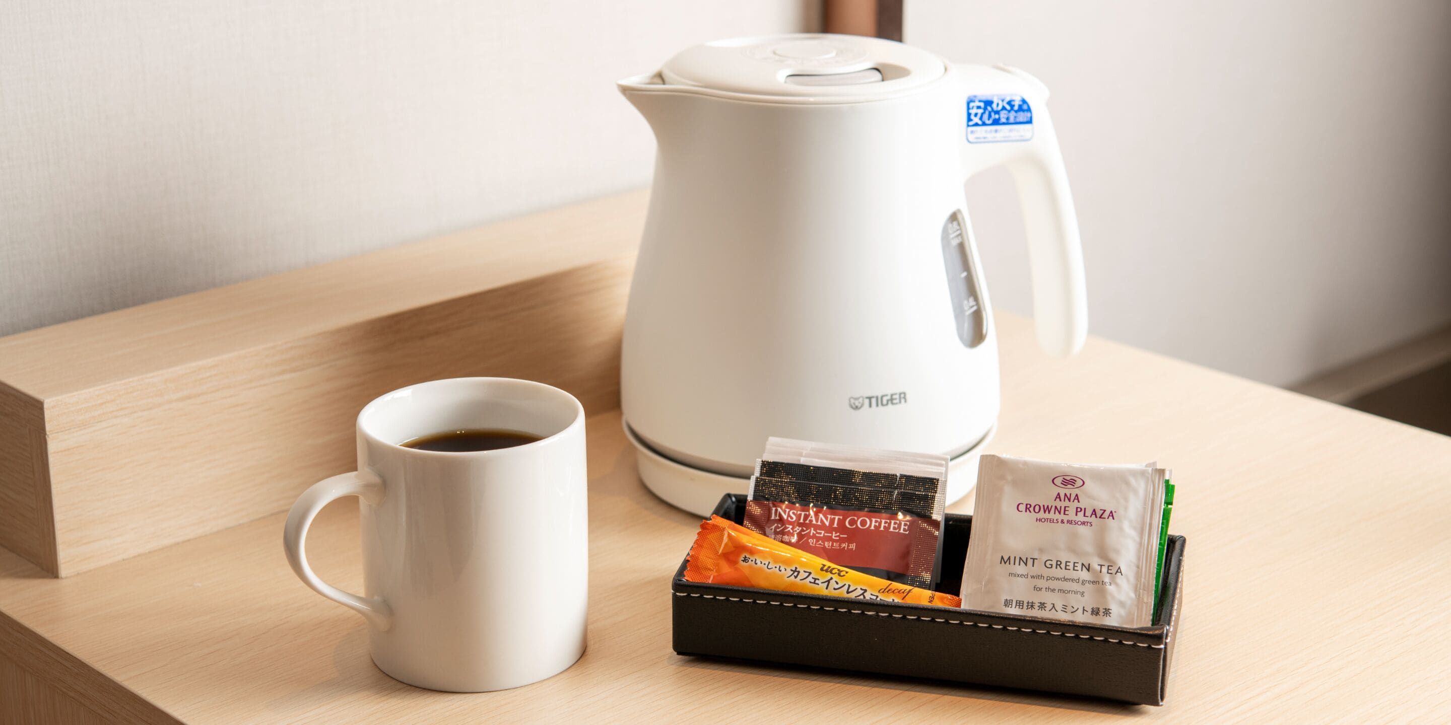 A white coffee maker, a white mug, and a black tray with instant coffee packets and a tea bag on a wooden surface.