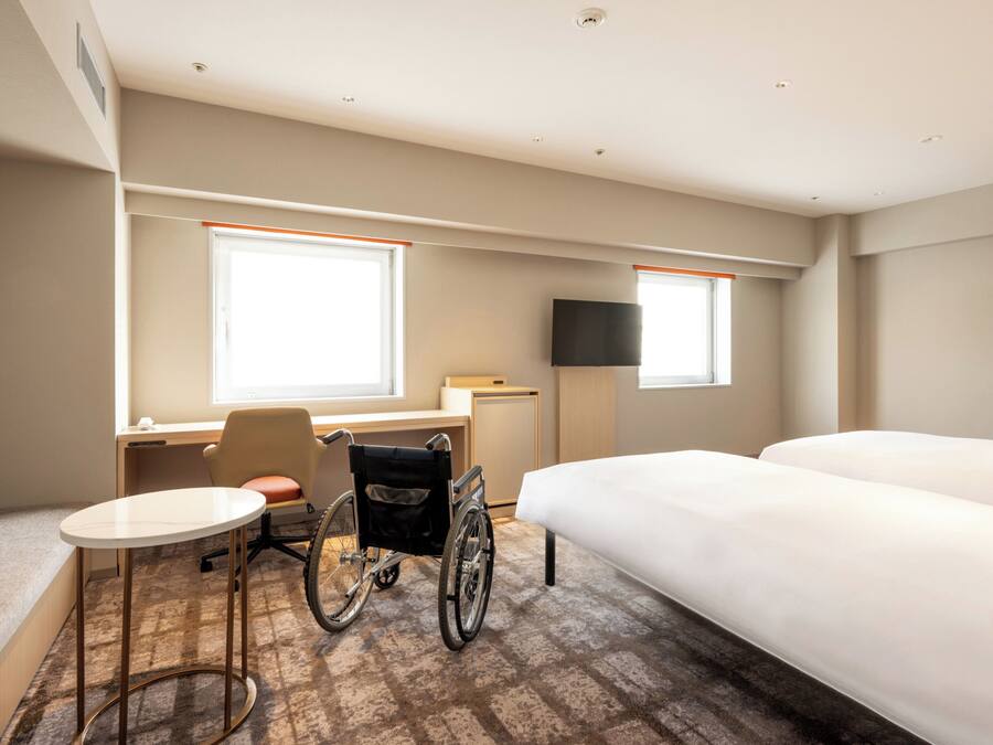 A hotel room with a bed, a desk, a wheelchair, a small table, and a TV.