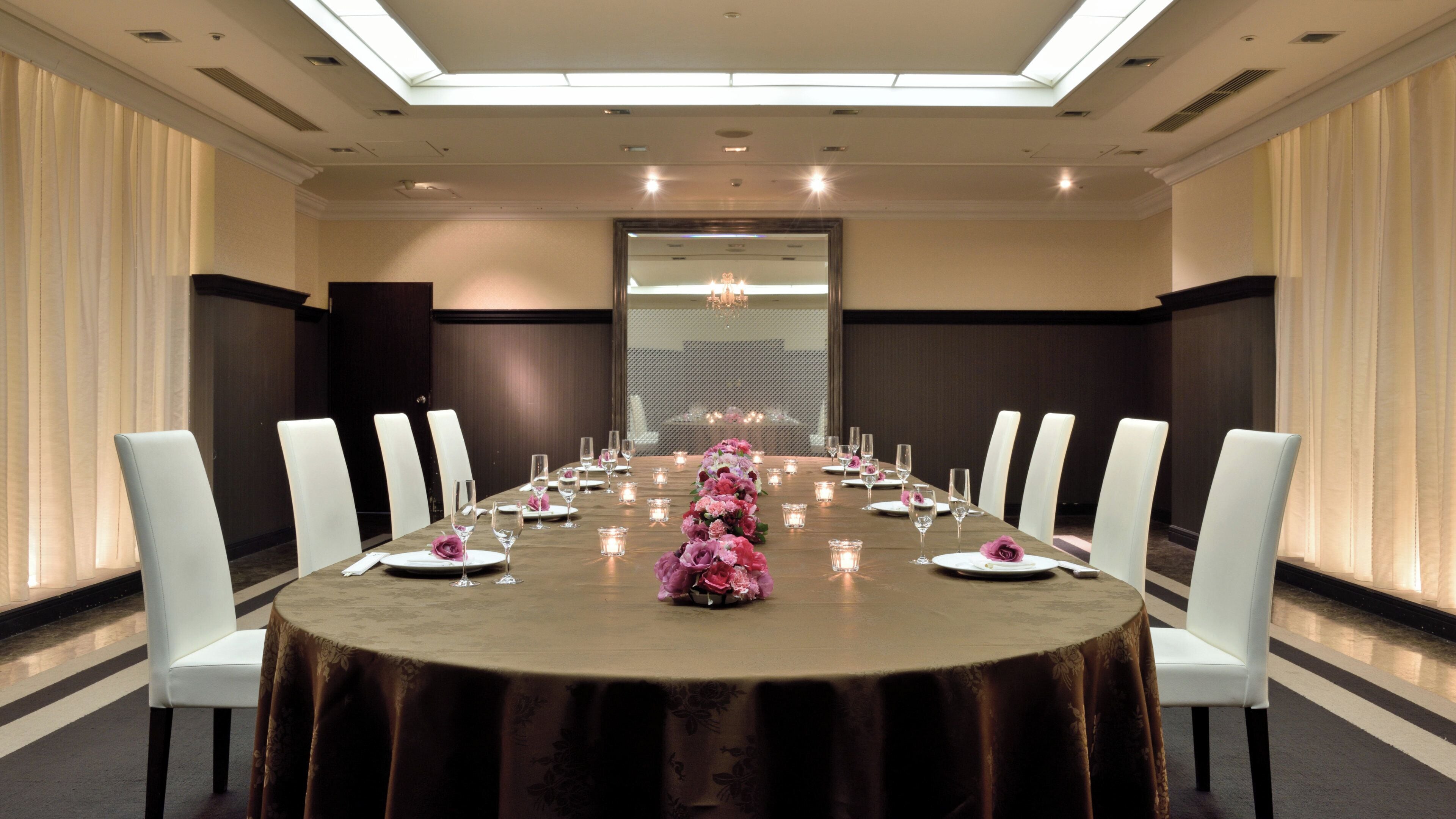A conference room with a long table set for a formal event, flanked by white chairs, and a large window in the background.