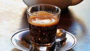 A clear glass of coffee with a spoon on a saucer, a teapot labeled "Maison Richard" in the background.