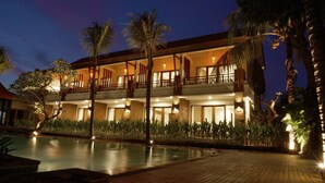 A two-story hotel with a pool, palm trees, and a well-lit exterior at dusk. - Arena Living (Denpasar)