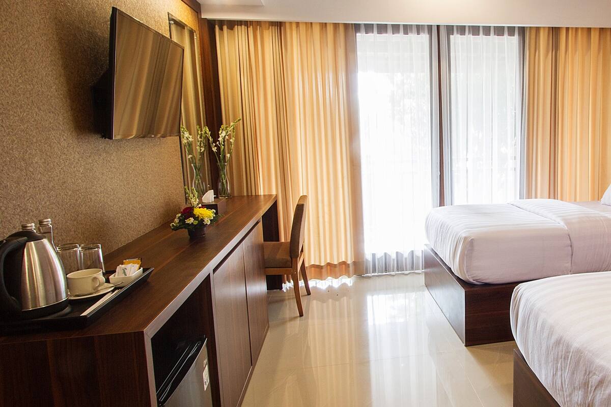 Habitación de hotel con dos camas, un escritorio con teléfono, una lámpara y vistas al exterior a través de amplias ventanas.