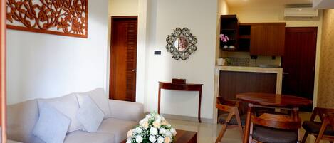 A living room with a sofa, coffee table, and a dining area with chairs and a table. There is a kitchen area with cabinets and an air conditioning unit.