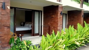 A hotel room with a bed, a small table, and a chair. There is a balcony with a view of the greenery outside. - Arena Living (Denpasar)