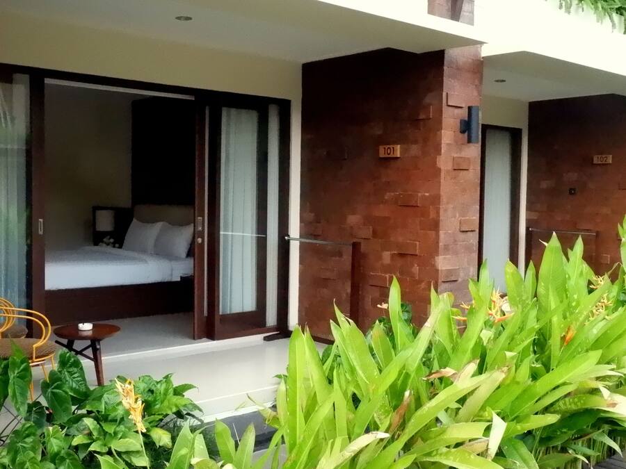A hotel room with a bed, a small table, and a chair. There is a balcony with a view of the greenery outside.