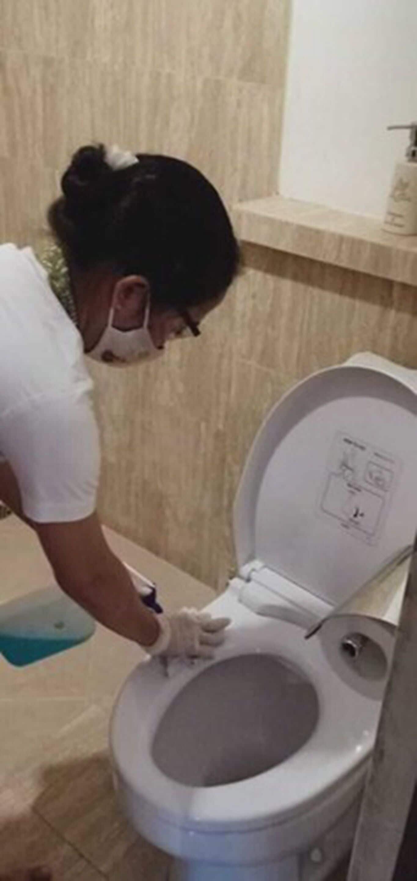 Uma pessoa limpando um vaso sanitário em um banheiro com chão de azulejos e uma prateleira acima.