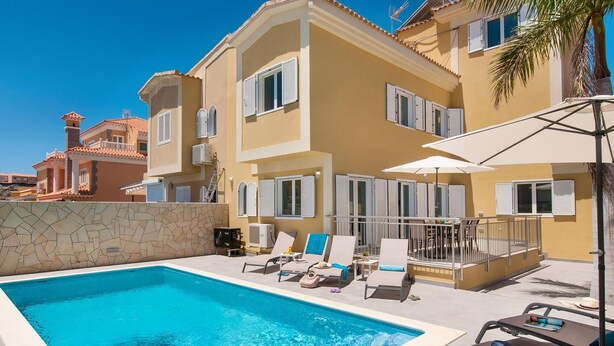 Uma área de piscina com espreguiçadeiras e guarda-sóis, uma parede de pedra e um edifício amarelo com persianas brancas.