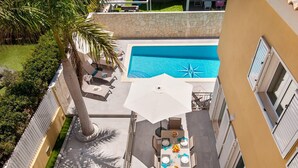 Uma área de piscina ao ar livre com mesa e cadeiras, cercada por um muro de pedra e palmeiras.