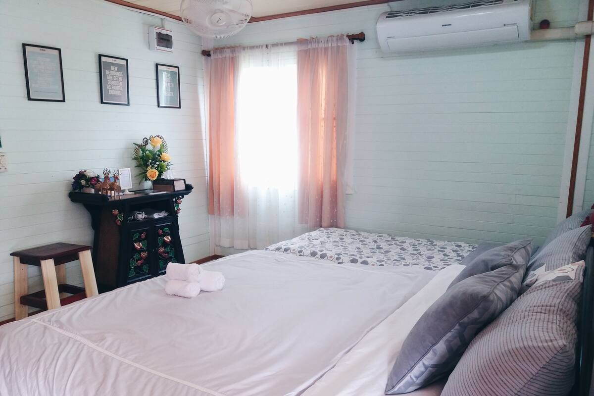 A neatly made bed with white bedding, a small wooden stool, a dresser with decorative items, and a window with sheer curtains.