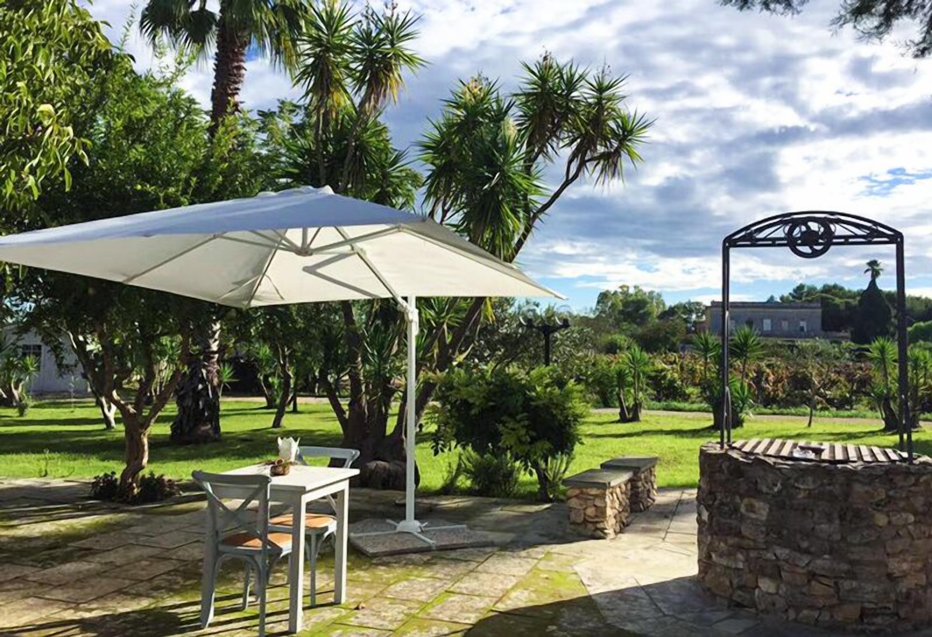 Un patio exterior con una mesa y sillas bajo un paraguas grande, rodeado de abundante vegetación y un muro de piedra.