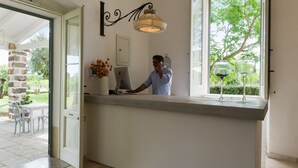 A reception desk with a computer, a decorative wall hanging, and a window with a view of greenery. - Dimora del Duca Masseria B&B (Monteroni di Lecce)