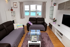 A living room with a sofa, a glass coffee table, a TV, and a window with a view of a body of water. - Cvita apartments (Trogir)