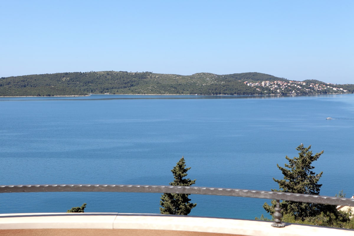 A coastal view with a clear sky, a calm sea, and a distant hill with buildings.