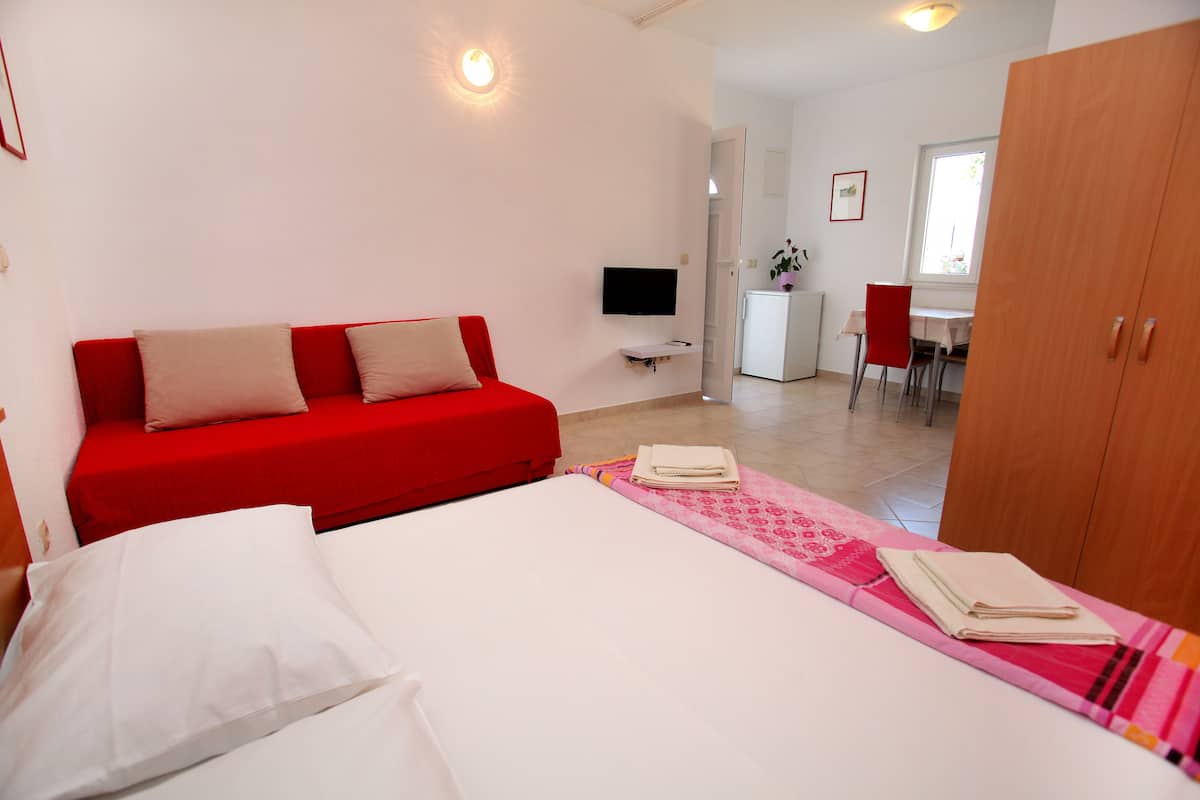A hotel room with a red sofa, a bed with white linens, a TV mounted on the wall, and a wooden wardrobe.