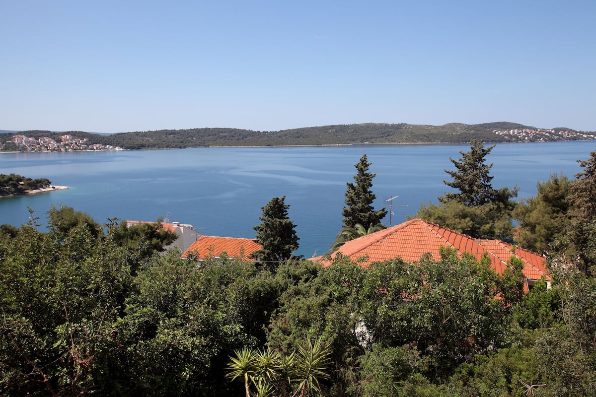 A coastal view with a clear blue sea, a red-roofed building, and dense greenery.